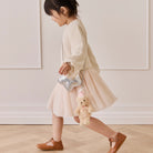 Plush tan bear keychain with pink party hat and bow, attached to silver star-shaped bag, worn by child in side view.