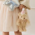 Fuzzy beige teddy bear keychain with pink party hat and bow, held by child in tulle skirt, lifestyle view