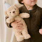 Soft beige plush teddy bear with green gingham party hat and bow, held by child in brown quilted jacket, lifestyle view