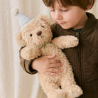 Soft beige plush teddy bear with green gingham party hat and bow, held by child in brown jacket, lifestyle side view