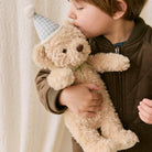 Soft beige plush teddy bear with green gingham party hat and bow, held by child in lifestyle shot