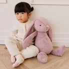 Soft pink plush bunny toy with long floppy ears and heart detail on foot, sitting beside child on wooden floor in lifestyle view