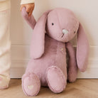 Soft pink plush bunny with long floppy ears and embroidered heart on foot, held by child in cream sweater, side view.