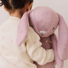 Soft pink plush bunny toy with long floppy ears and silver nose, held by child in cream floral sweater, back view.