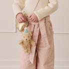 Fuzzy tan teddy bear keychain with blue party hat and bow, attached to gold bag, worn by child in cream sweater and pink corduroy pants, lifestyle view