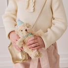 Plush beige teddy bear keychain with blue party hat and bow, held by child in cream ribbed sweater, lifestyle front view
