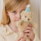 Soft beige plush bear keychain with light blue party hat and matching bow, held by child in lifestyle close-up view