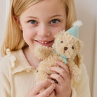 Soft beige plush bear keychain with light blue party hat and bow, held by child in cream ruffled top, front view