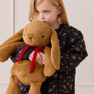 Brown plush bunny toy with long floppy ears and red ribbon, held by child in floral outfit, lifestyle view