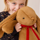 Brown plush bunny with long ears and red velvet bow, held by child in floral dress, lifestyle close-up view